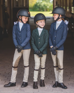 three children in a barn wearing full equestrian gear smiling at one another
